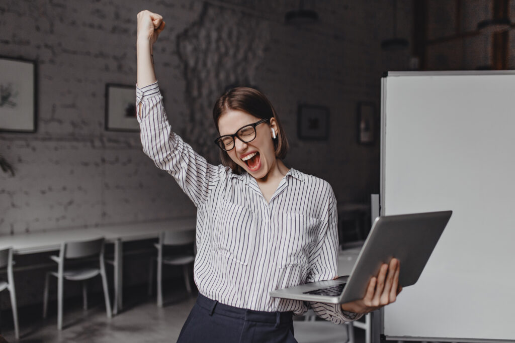 Business woman with laptop in hand is happy with success. Portrait of woman in glasses and striped blouse enthusiastically screaming and making winning gesture.