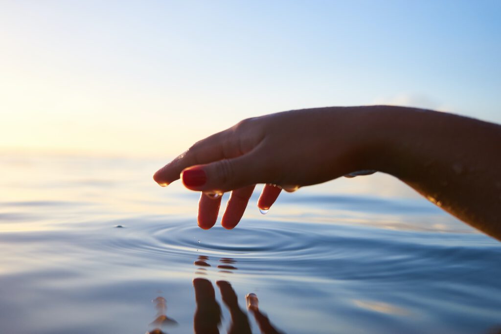 יד של אישה נוגעת במים בזמן זריחה, מייצרת אדוות והשתקפות