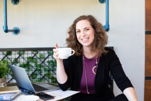 אישה עצמאית יושבת מול שולחן עם מחשב וכוס קפה ביד צילומי תדמית עצמאיות מהבטן קהילה עסקית