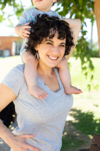 אישה עצמאית עם ילד על הכתפיים. היא לובשת חולצה אפורה וברקע ירוק ומדשאות. צילומי תדמית מהבטן