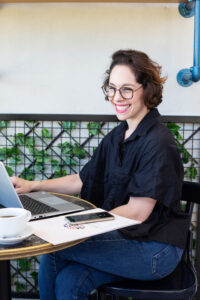 אישה עם משקפיים יושבת ליד שולחן. עליו מונח מחשב, מחברת וכוס קפה. ברקע קיר לבן, בתחתיתו רשת שחורה עם עלים ירוקים. צילומי תדמית מהבטן