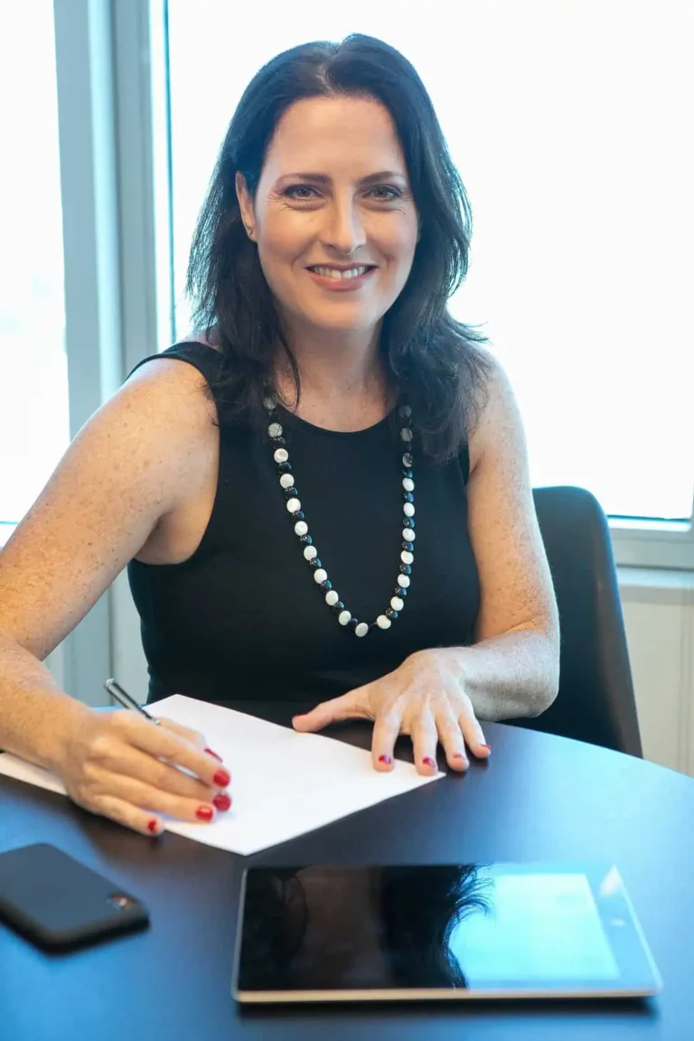 אורית בש סרוסי, אישה עצמאית יושבת ליד שולחן עבודה. היא לובשת חולצה שחורה, עונדת שרשרת פנינים, שיערה מפוזר ומחייכת. מאחוריה חלון ועל השולחן מונח טאבלט, נייד ובלוק כתיבה