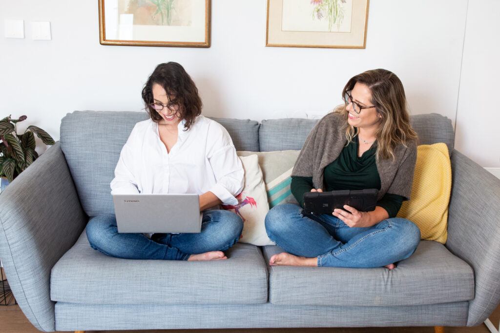 שתי נשים עובדות על ספה, אחת עם סוודר ירוק וג'ינס והשנייה עם חולצה לבנה וג'ינס. מאחוריהן תמונות של פרחים והן מחייכות ונהנות מהעבודה בבית